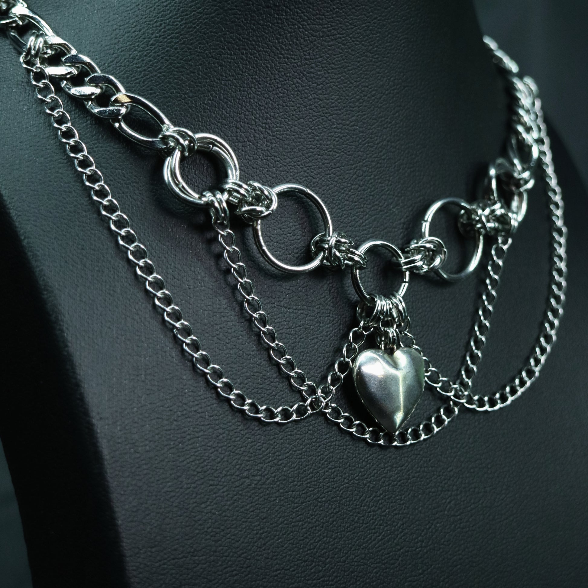 Angled view of a handmade stainless steel choker featuring circular rings, layered chain drapes, and a central faceted heart charm.