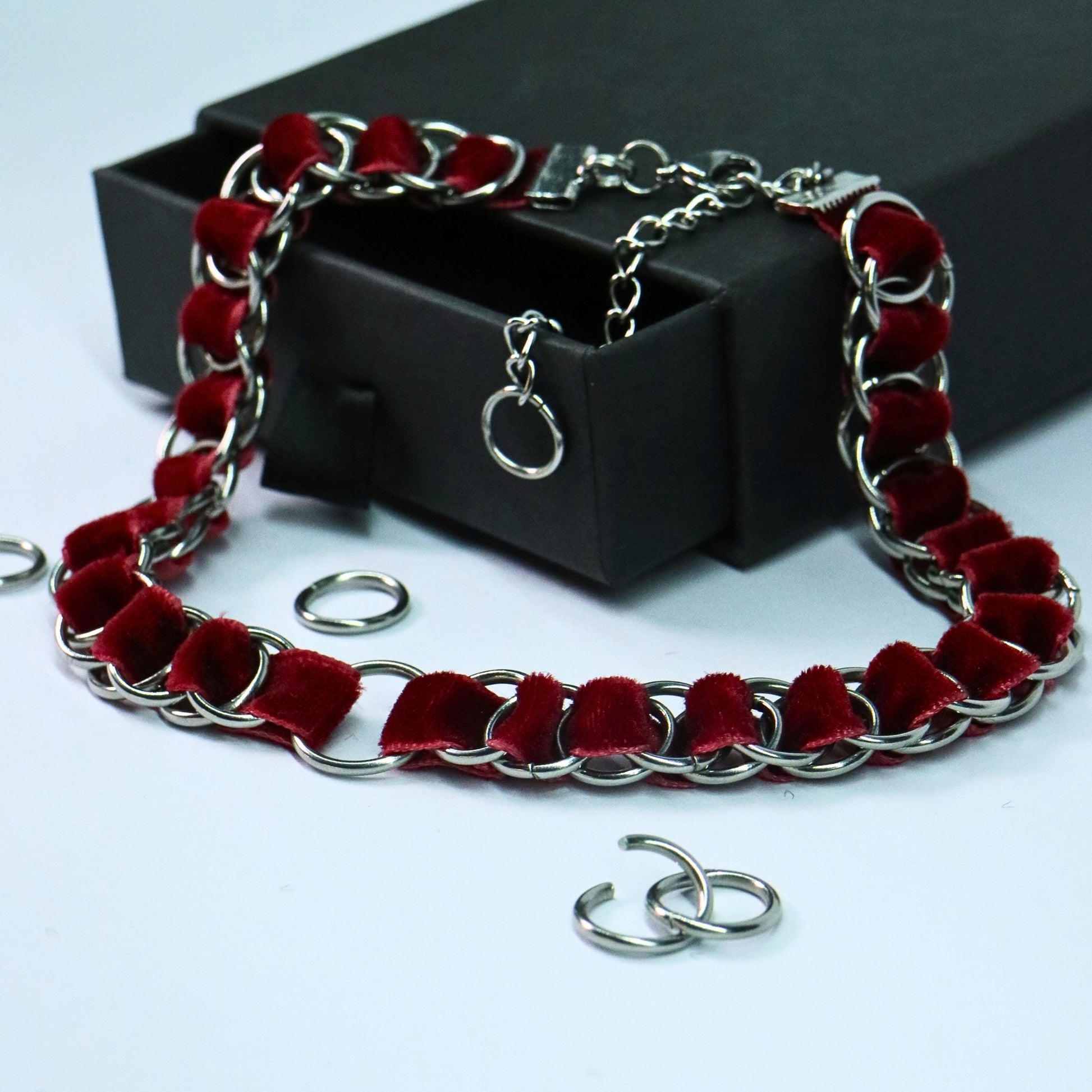 Blood red velvet ribbon choker interlaced with stainless steel rings and an O-ring center, shown next to a black gift box.
