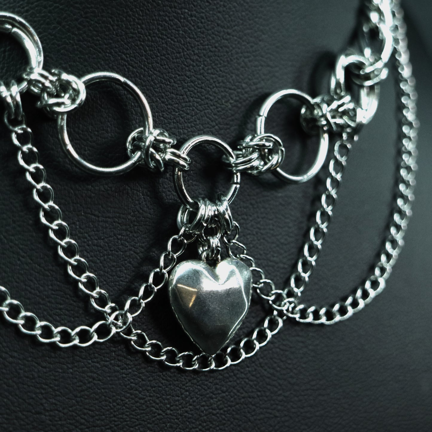 Close-up of a layered stainless steel choker with interlinked rings, draped chains, and a faceted rhodium-plated heart pendant.