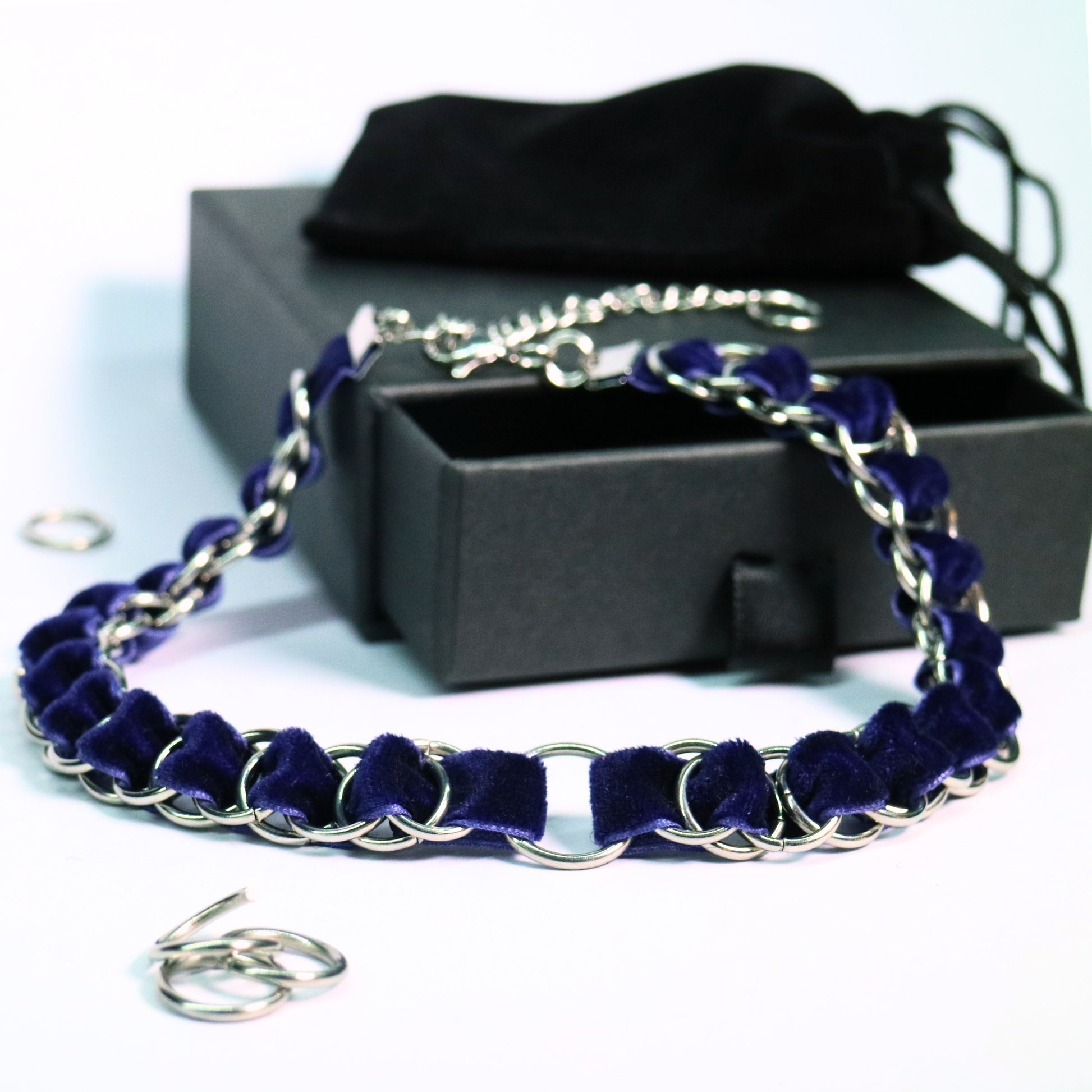 Deep blue velvet ribbon choker woven through stainless steel rings with an O-ring centerpiece, displayed beside a black gift box.