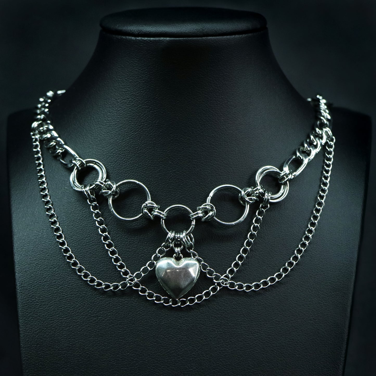 Front view of a layered stainless steel choker with ring links, double-chain draping, and a polished faceted heart pendant.