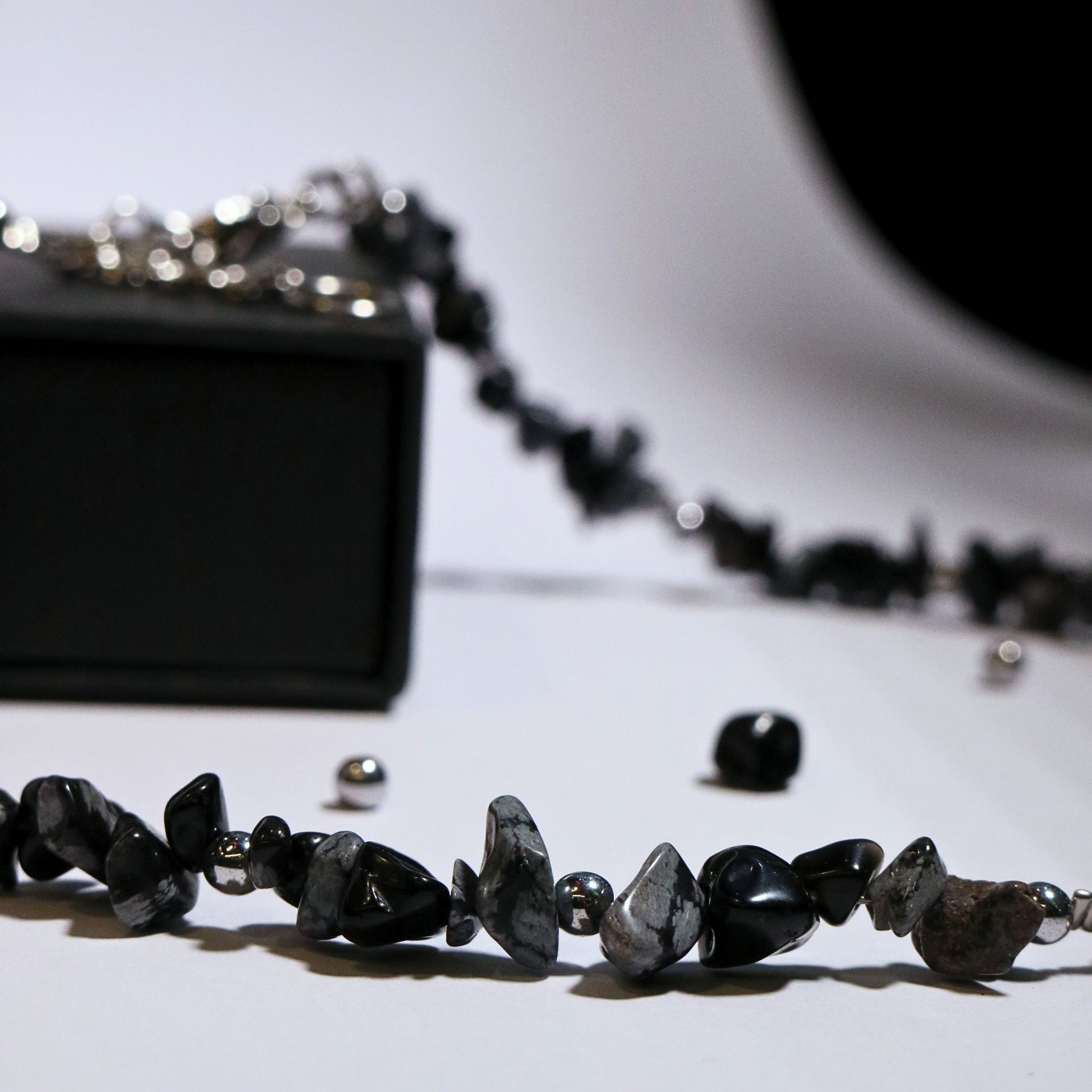 Side view of a handmade natural-stone necklace in black and grey tones draped over a black box, showing bead texture and shine.