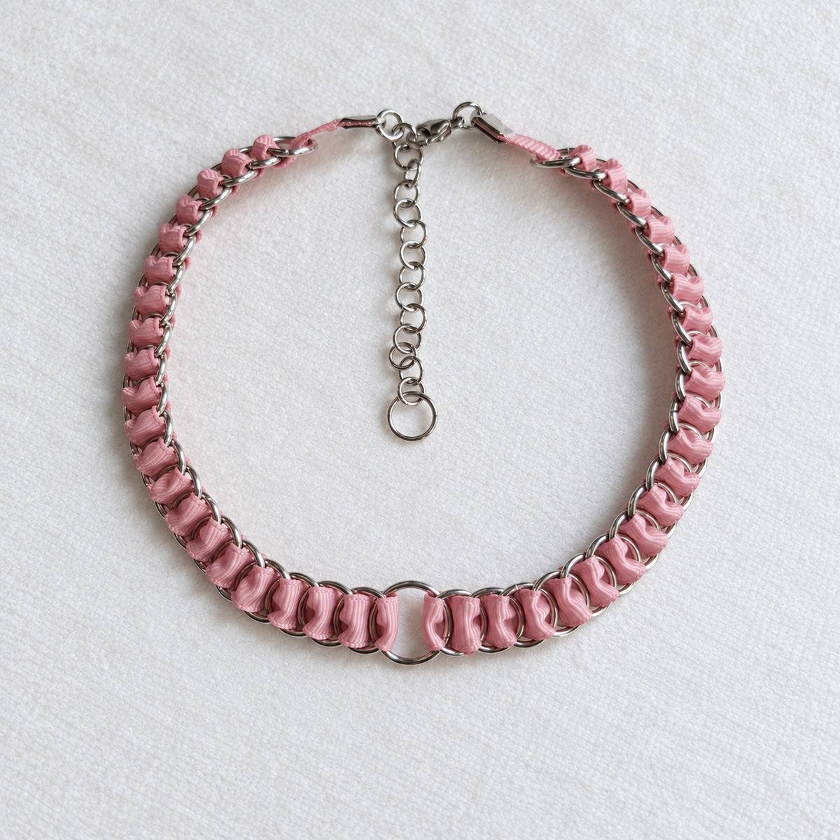 Overhead view of the LOOM dusty pink ribbon chainmail choker with stainless steel central O-ring, clasp and extension chain on white fabric.