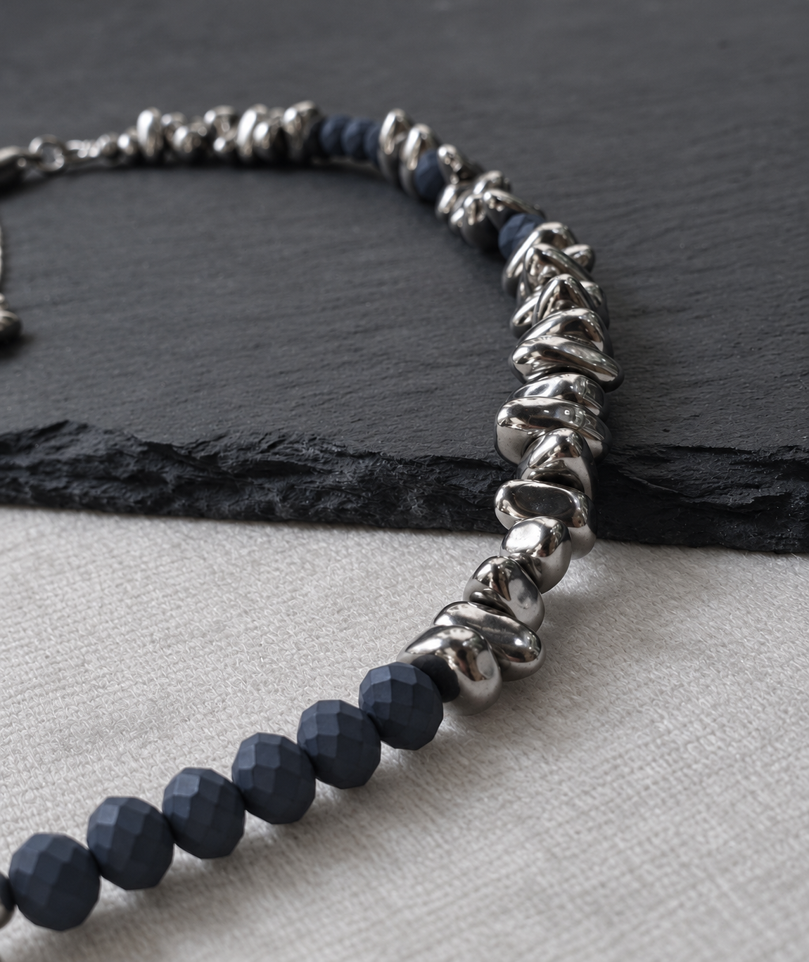 Detail of NIGHTMARK's irregular polished silver hematite beads alongside matte midnight blue crystal beads on slate and white fabric.
