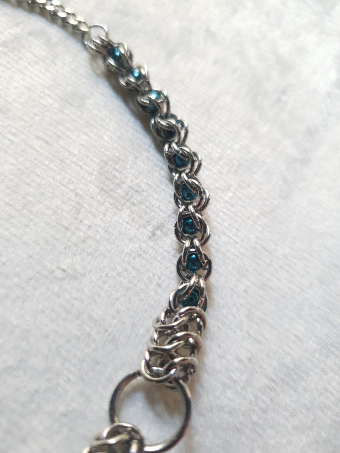 A close-up of a silver-toned chain bracelet featuring small teal blue heart-shaped inlays, laid on a textured white fabric background.