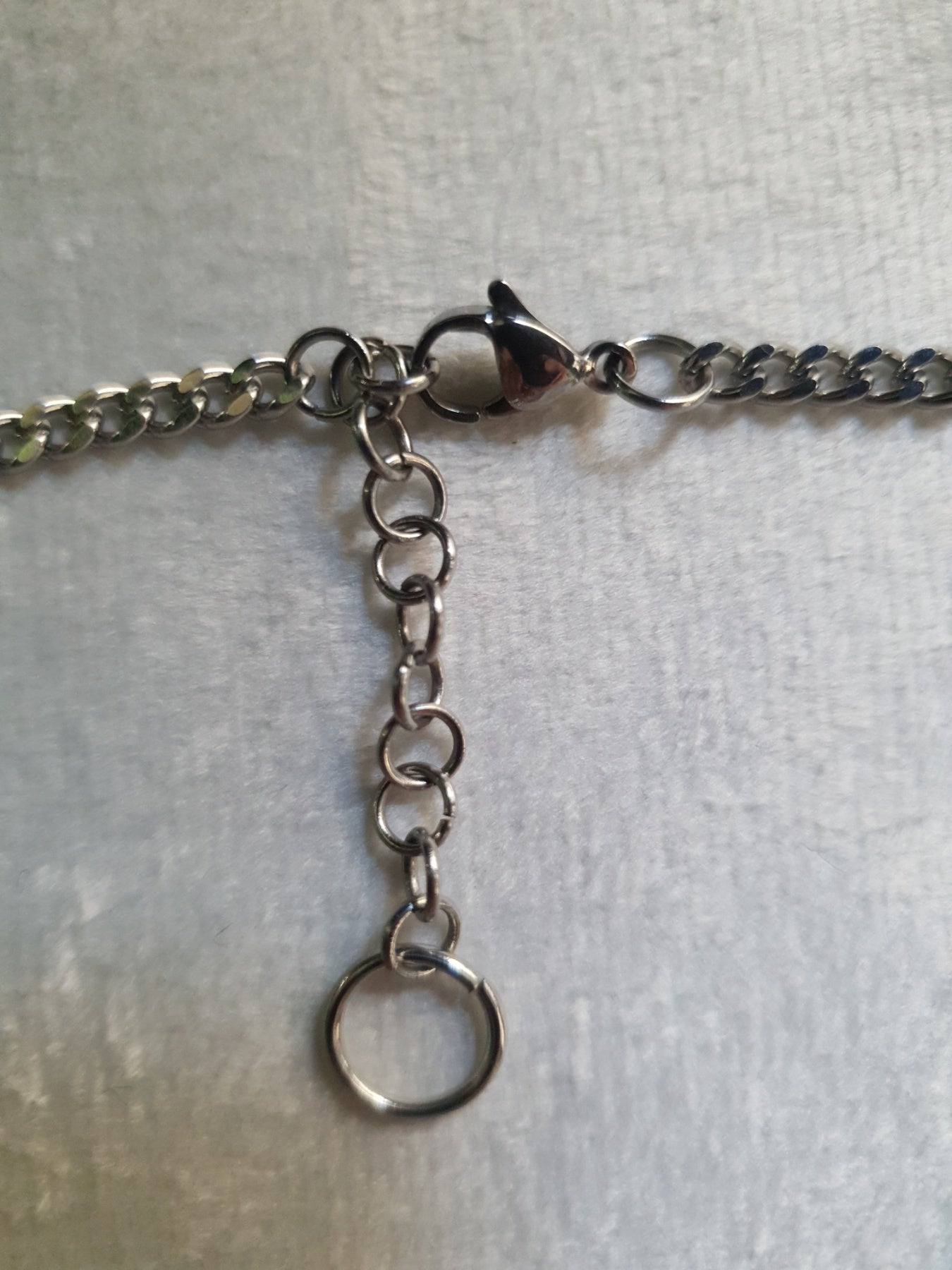Close-up of a silver-toned chain necklace with a lobster clasp, extension chain, and round ring ending, laid on a light gray textured surface.